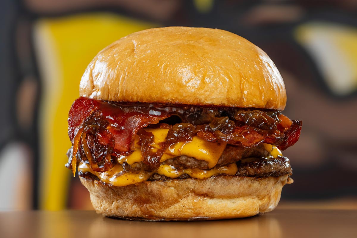 Hamburguesa doble con queso derretido bacon crujiente y cebollas caramelizadas sobre pan tostado en un restaurante cálido con fondo desenfocado transmitiendo una sensación indulgente